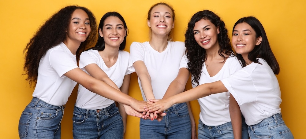 Five Diverse Women Holding Hands Standing On Yellow Studio Background