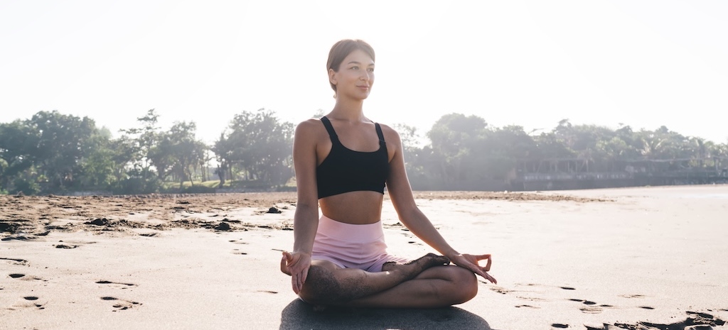 Young woman sitting in lotus pose enjoying yoga practice on the beach in San Diego in trauma treatment.