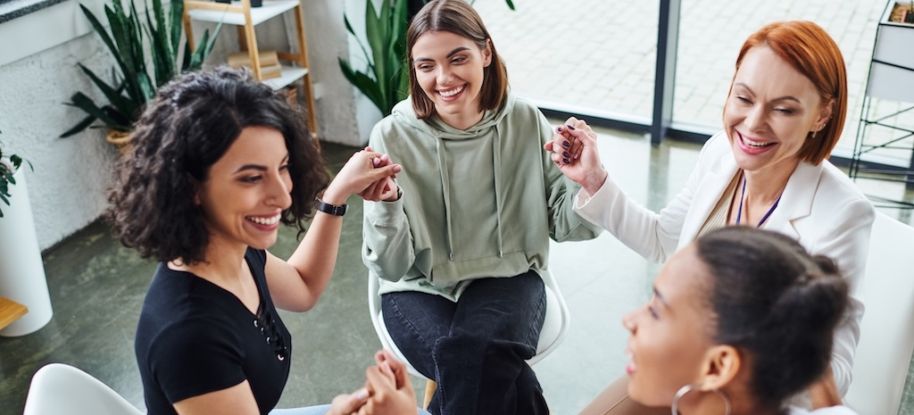 cheerful multicultural female friends holding hands and communicating during group therapy