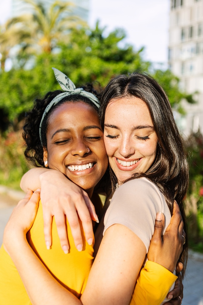 Vertical portrait of candid happy best women friends embracing outdoors. Close up view of two diverse girls hugging each other with closed eyes smiling.